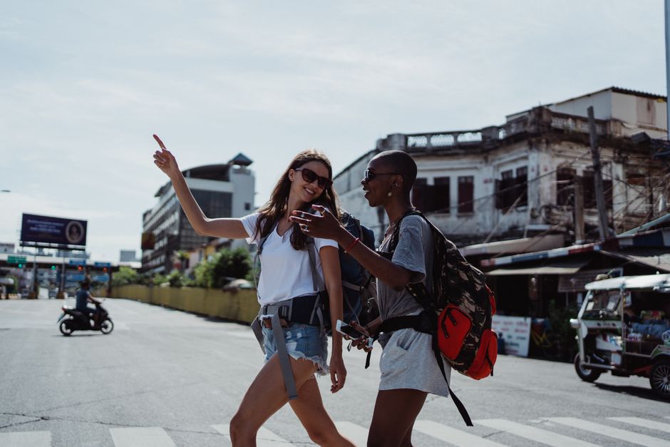 Two women crossing a busy street, exploring a vibrant city while wearing backpacks, capturing the essence of urban travel.