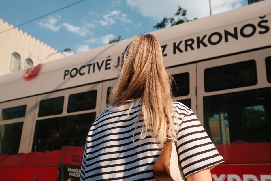 A woman with blonde hair in a striped shirt stands near a city tram on a sunny day.