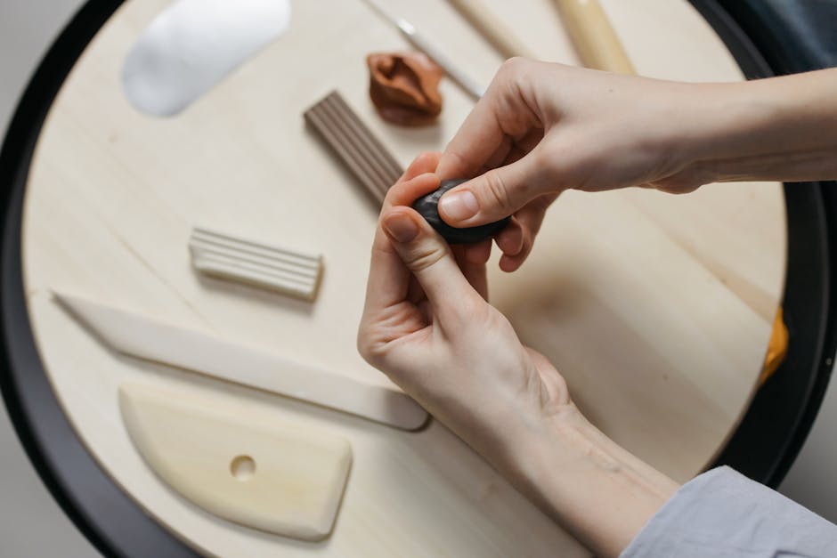 Hands shaping clay with various tools on a wooden surface, showcasing artistic creativity.
