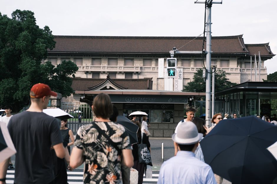 A lively urban scene featuring people crossing in front of a historical building on a cloudy day.