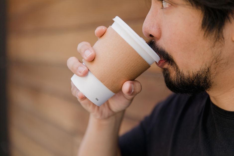 Close-up of bearded man drinking coffee from a disposable cup. Soft, neutral tones with wooden background.