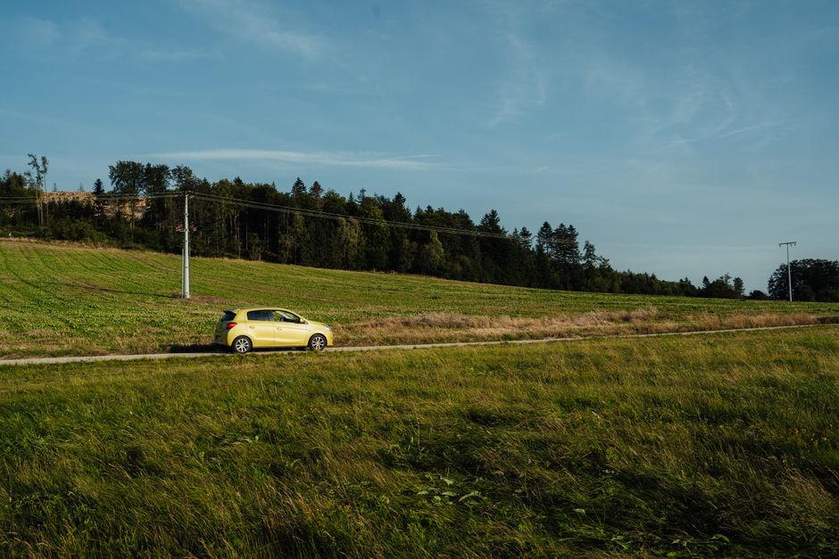 A yellow car travels down a country road through grassy fields and forests under a clear sky.