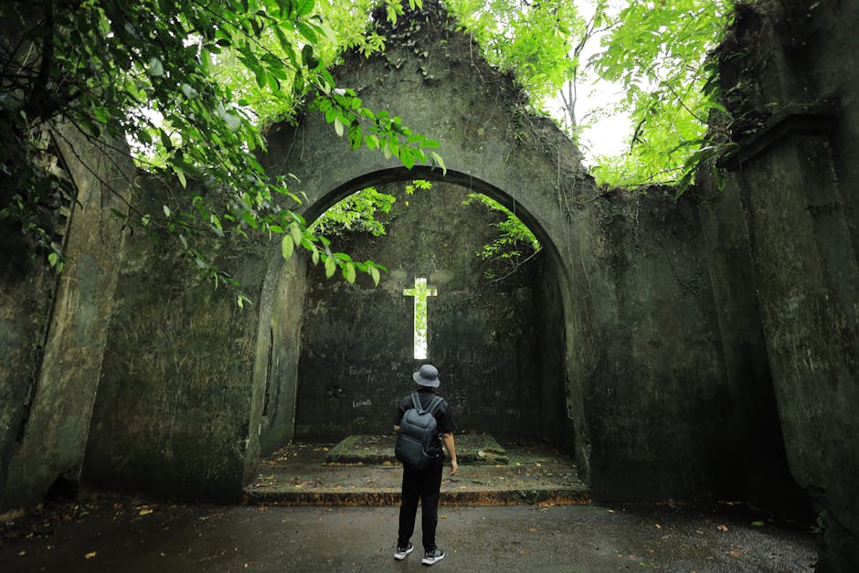 A traveler stands inside a moss-covered, ruined chapel with a stone cross in Việt Nam's lush greenery.