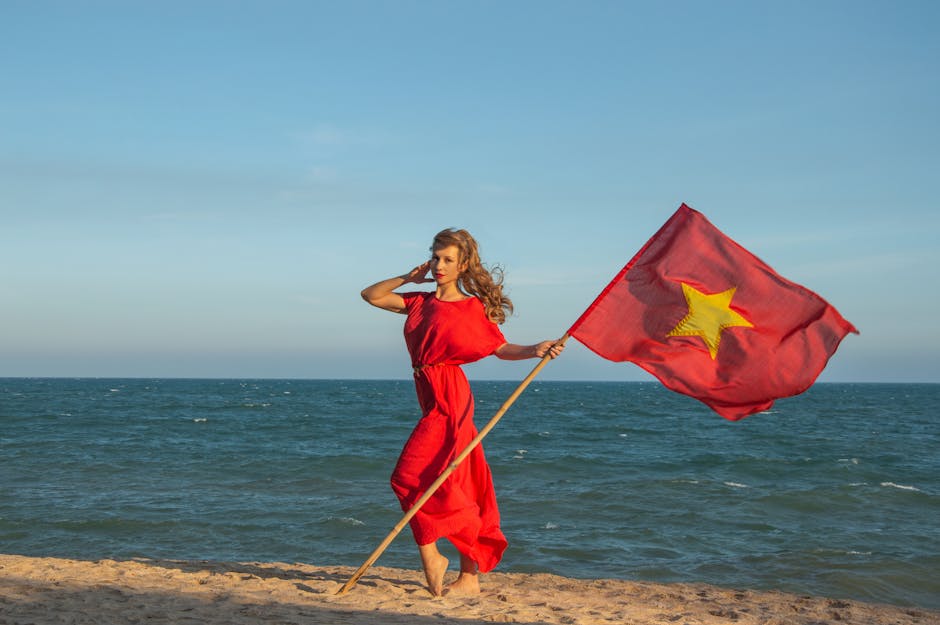 A woman in a red dress stands on a beach holding the Vietnamese flag against a clear sky.