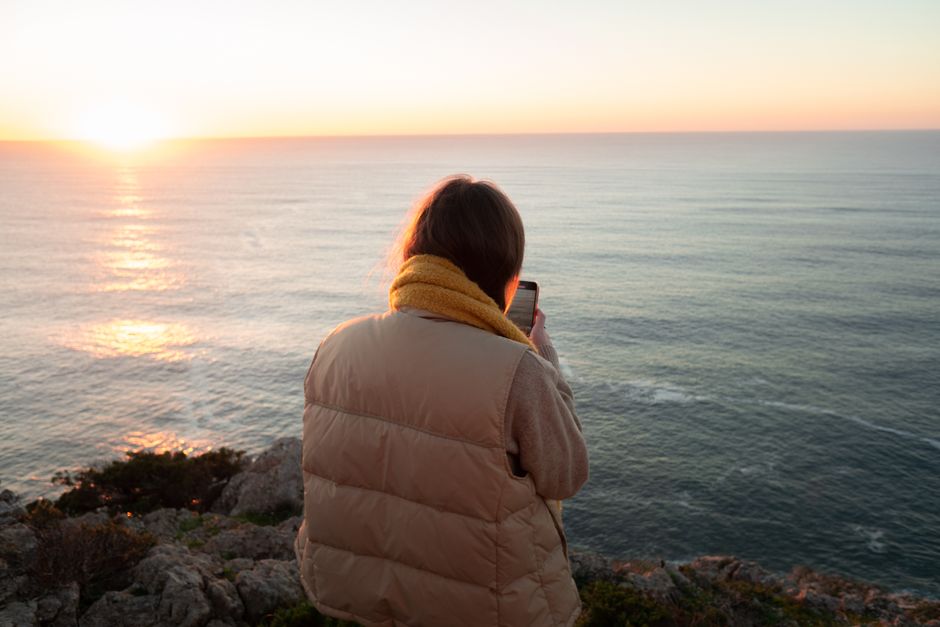 A woman in a brown jacket and yellow scarf takes a photo of the ocean at sunset.