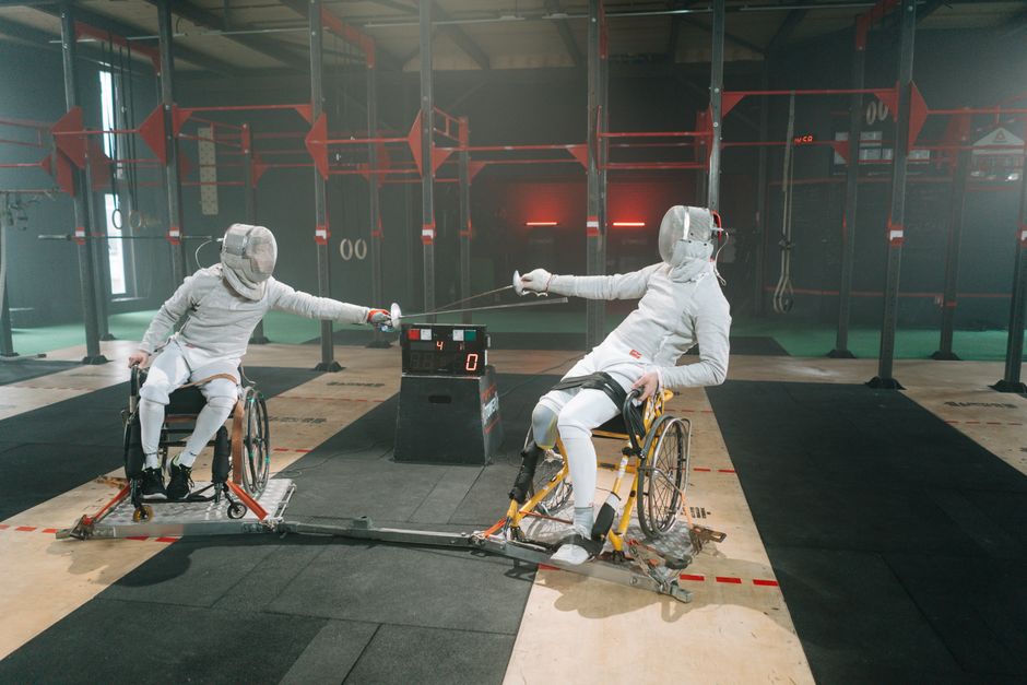 Two wheelchair fencers engaging in a competitive indoor fencing match.
