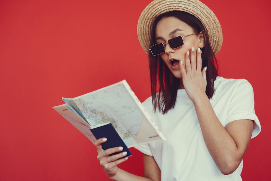Surprised young woman with long dark hair in stylish sunglasses looking at map and touching face while checking travel route on red background